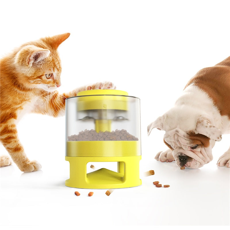 Orange tabby cat interacting with a yellow Gravity Catapult Puzzle Feeder - Interactive IQ Training & Slow Feed Station while a bulldog eats kibble from the floor nearby.