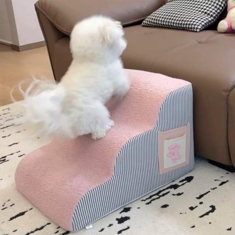 Small white dog climbing pink and striped Orthopedic Curved Dog Ramp - High-Density Foam Pet Stairs for Small & Senior Pets - IVDD Safe placed next to a sofa.