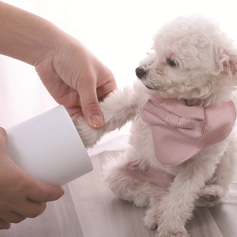 Small white dog wearing pink bow tie having its paw cleaned using the Portable Dog Foot Washer - Professional Muddy Paw Cleaning Cup held by a person’s hand against a light background.