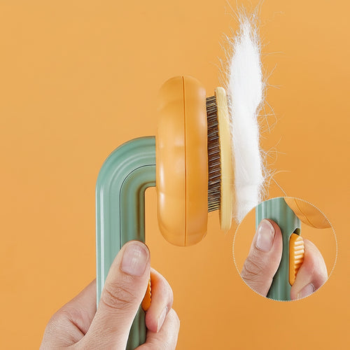 Hand holding Boutique Pumpkin Self-Cleaning Brush - Aesthetic Grooming & Deshedding Comb with white pet hair being released from bristles by pressing a thumb button on a green and orange handle against a solid orange background.