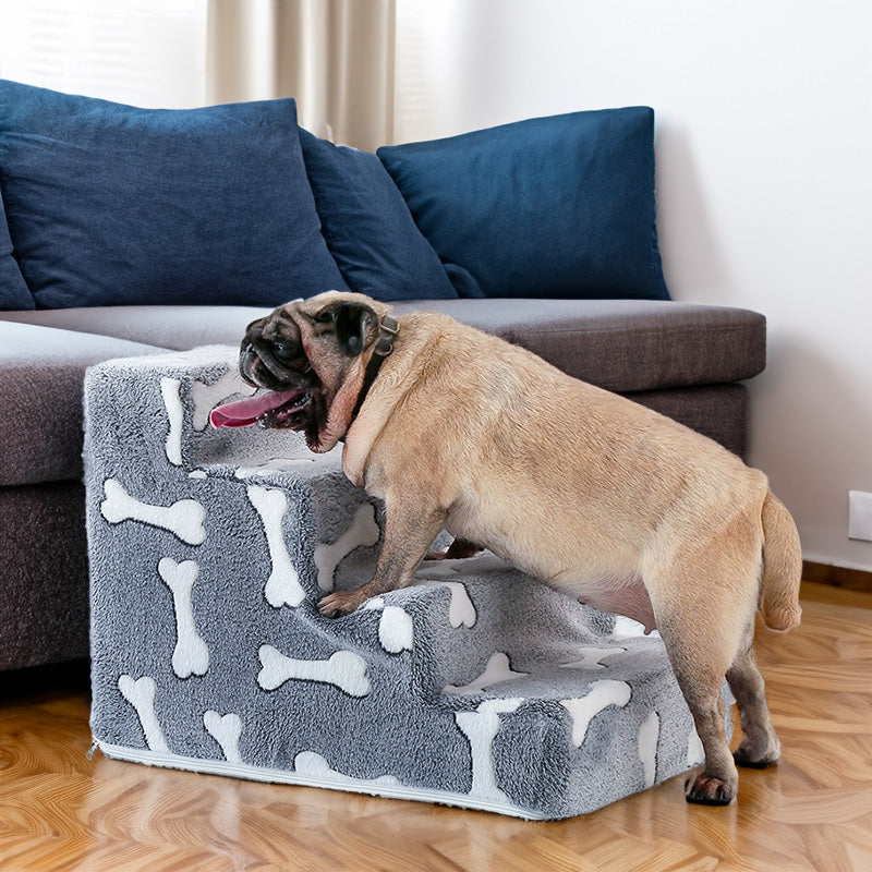 A pug using Tiered High-Density Foam Pet Stairs - Nonslip Pet Steps for High Beds & Sofas - Easy-Access Joint Support with a soft gray bone pattern next to a sofa with blue cushions.