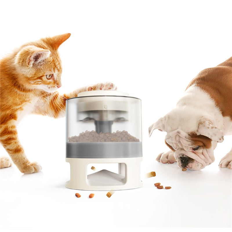 Orange tabby cat pawing at the Gravity Catapult Puzzle Feeder - Interactive IQ Training & Slow Feed Station while a bulldog eats scattered kibble nearby on a white background