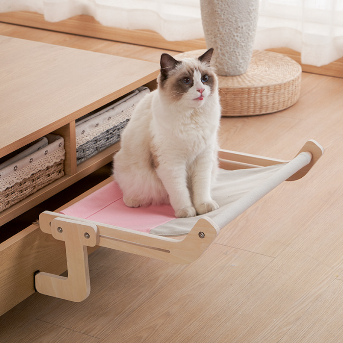 A fluffy cat sitting on a pink fabric shelf mounted on a wooden low cabinet, showcasing the No-Suction Wooden Cat Window Perch - Heavy-Duty Adjustable Hook-On Shelf & Safety Sun Lounger in a bright room.