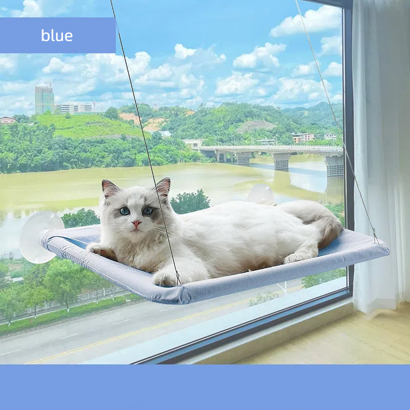 White cat resting on a blue Foldable Cat Window Hammock - Space-Saving Suction Cup Perch & Elevated Sunbed attached to a large window overlooking a river and greenery outside.