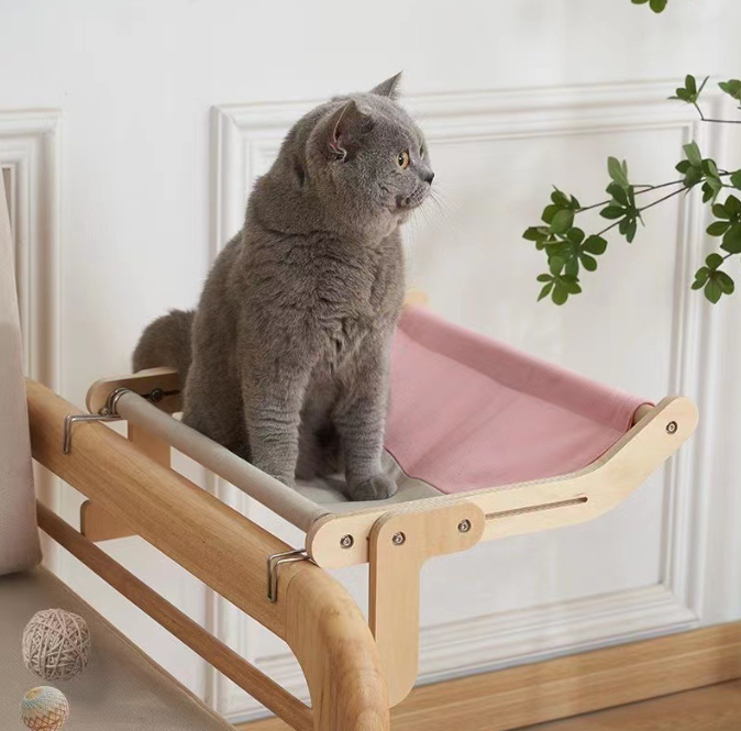 A gray cat sits comfortably on a pink fabric platform attached to a wooden frame, showcasing the No-Suction Wooden Cat Window Perch - Heavy-Duty Adjustable Hook-On Shelf & Safety Sun Lounger.