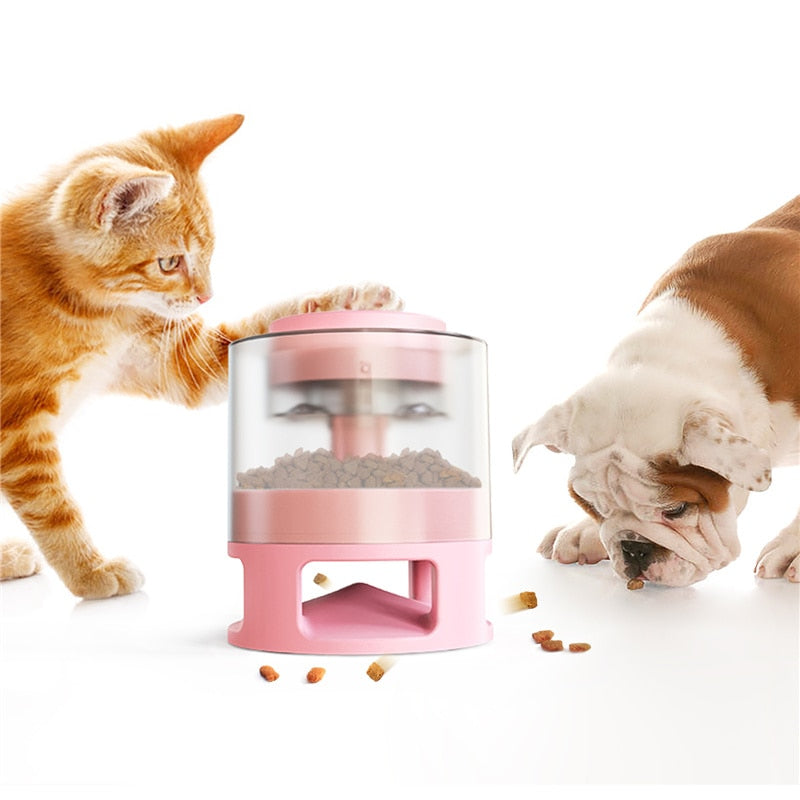 Orange tabby cat interacting with a pink Gravity Catapult Puzzle Feeder - Interactive IQ Training & Slow Feed Station while a bulldog eats kibble from the floor nearby.