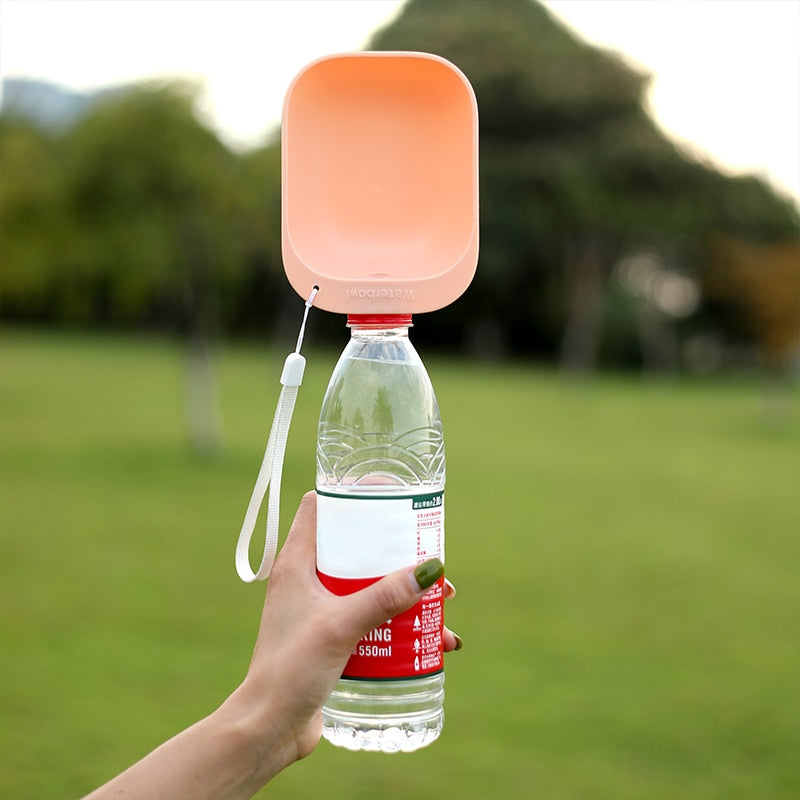 Hand holding a clear plastic bottle fitted with a Universal Pet Hydration Adapter – Portable Bottle Attachment & Travel Drinking Scoop in peach, outdoors on a blurred green lawn background.